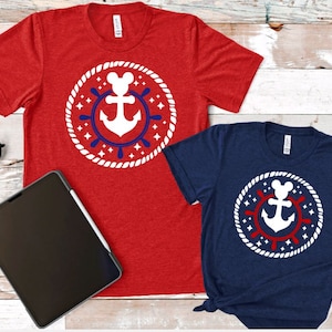 May include: Two short-sleeved t-shirts, one red and one navy blue, with a white anchor design in a circular frame. The frame has a rope border, a red and blue wheel, and star accents. The shirts are on a wooden surface.