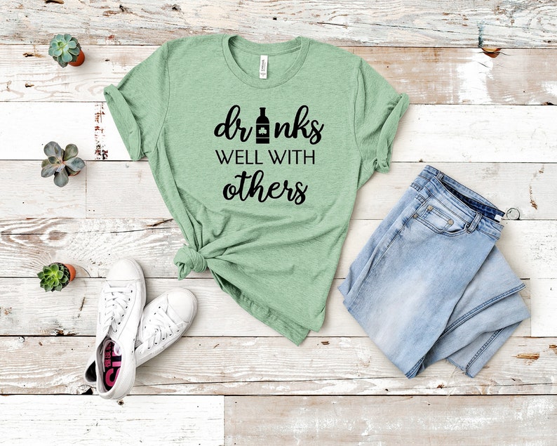 May include: A light green t-shirt with the text "drinks well with others" in black. The shirt is paired with light blue jeans and white sneakers. Small potted succulents are arranged around the clothing.