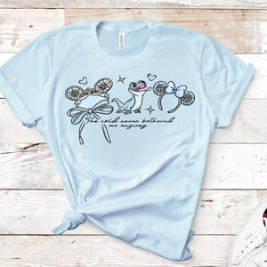 Może przedstawiać: Jasnoniebieski t-shirt z nadrukiem graficznym przedstawiającym małą jaszczurkę, dwie pary mysich uszu z kokardkami oraz napis "The cold never bothered me anyway." Wz&oacute;r zawiera małe akcenty w kształcie płatk&oacute;w śniegu i gwiazdek w stylu liniowym.