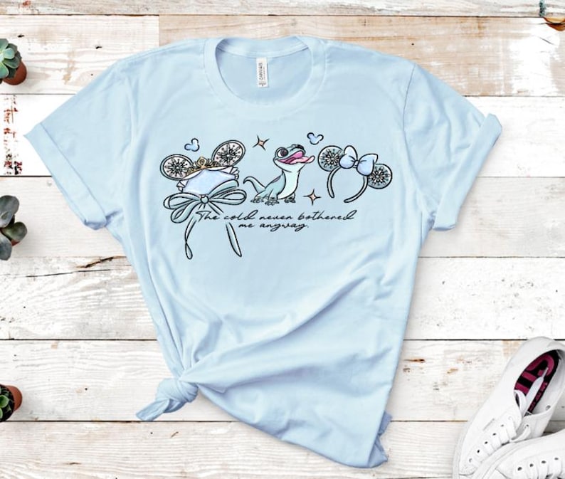 Może przedstawiać: Jasnoniebieski t-shirt z nadrukiem graficznym przedstawiającym małą jaszczurkę, dwie pary mysich uszu z kokardkami oraz napis "The cold never bothered me anyway." Wz&oacute;r zawiera małe akcenty w kształcie płatk&oacute;w śniegu i gwiazdek w stylu liniowym.