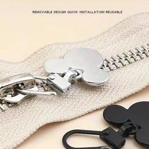 Op de afbeelding: Een close-up van een rits met een zilveren schuif en een Mickey Mouse-vormige rits trek. De rits is beige en heeft een witte achtergrond. De rits trek is zilver en heeft een zwart Mickey Mouse hoofd.