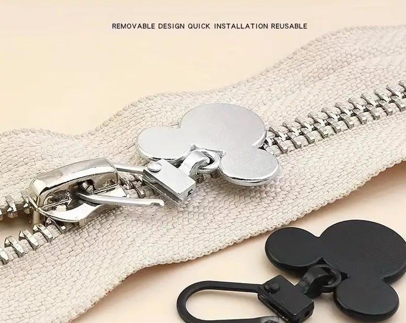 Op de afbeelding: Een close-up van een rits met een zilveren schuif en een Mickey Mouse-vormige rits trek. De rits is beige en heeft een witte achtergrond. De rits trek is zilver en heeft een zwart Mickey Mouse hoofd.