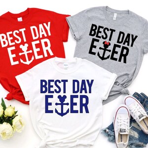 May include: Three short-sleeved t-shirts in red, white, and gray, each with the text "BEST DAY EVER" and an anchor design. The anchor design incorporates a Mickey Mouse head. A pair of white sneakers and denim shorts are also visible.