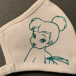 Könnte beinhalten: Weiße Gesichtsmaske mit einer grünen Glitzerkontur von Tinkerbell. Die Maske besteht aus weißem Stoff mit einer genähten Bordüre und Ohrenschlaufen. Das Design ist eine skurrile Illustration der Disney-Figur.