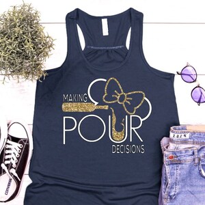 May include: Navy blue tank top with the phrase "MAKING POUR DECISIONS" in white and gold glitter. The design includes a wine bottle and bow. The tank top is paired with jeans, sunglasses, and sneakers.