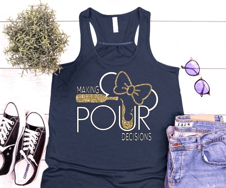 May include: Navy blue tank top with the phrase "MAKING POUR DECISIONS" in white and gold glitter. The design includes a wine bottle and bow. The tank top is paired with jeans, sunglasses, and sneakers.