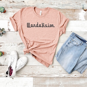 May include: A peach-colored t-shirt with the text "WandaVision" in black. The shirt is paired with light blue jeans and white sneakers. Small potted succulents are arranged around the clothing.