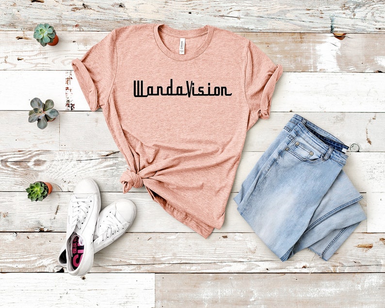 May include: A peach-colored t-shirt with the text "WandaVision" in black. The shirt is paired with light blue jeans and white sneakers. Small potted succulents are arranged around the clothing.