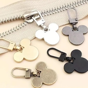 Może przedstawiać: Pięć suwaków do zamków błyskawicznych z motywem głowy Myszki Miki. Suwaki są w kolorze srebrnym, złotym, czarnym i brązowym.