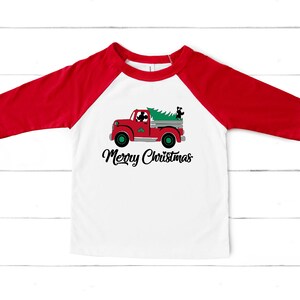 May include: A white and red raglan-style top featuring a red truck carrying a Christmas tree. The truck has a black dog in the driver's seat and another in the back. The words "Merry Christmas" are written in black script below the truck.