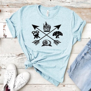 May include: A light blue t-shirt with a black graphic design featuring a castle, Mickey Mouse head, a tree, and a sphere with a crosshatch pattern. The graphic is surrounded by four black arrows pointing in different directions.