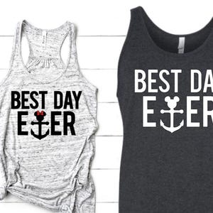 May include: Two vest tops, one light grey and one dark grey, each with the text "BEST DAY EVER" in white. The word "EVER" features an anchor design. The light grey vest top has a red bow in the design.