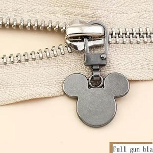 Op de afbeelding: Een zwarte metalen rits met een Mickey Mouse-ontwerp. De rits is bevestigd aan een zilveren rits.