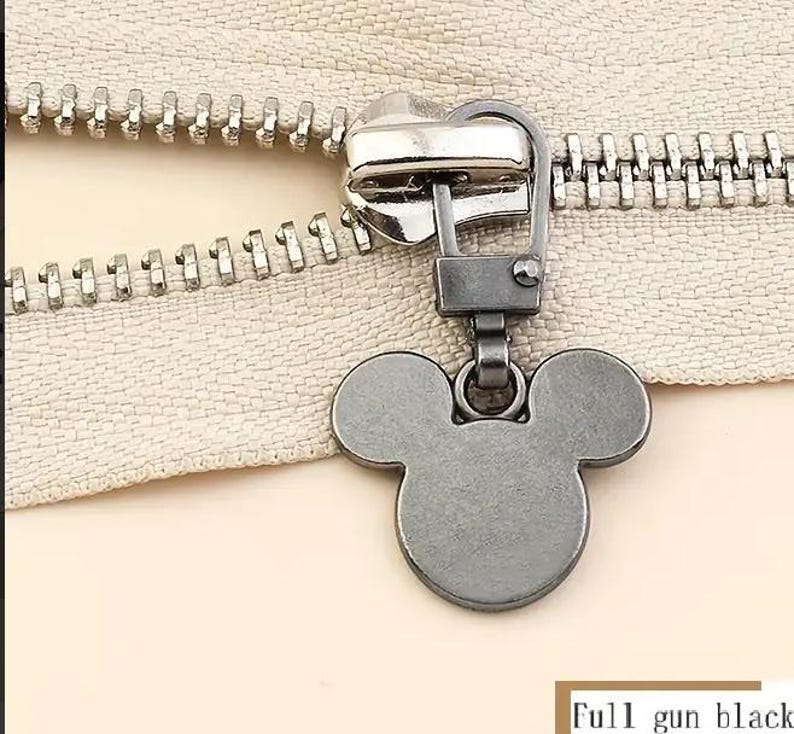 Op de afbeelding: Een zwarte metalen rits met een Mickey Mouse-ontwerp. De rits is bevestigd aan een zilveren rits.