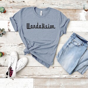 May include: A light blue t-shirt with the word "WandaVision" printed in black. The shirt is paired with light blue jeans and white sneakers. Small potted succulents are arranged around the clothing.