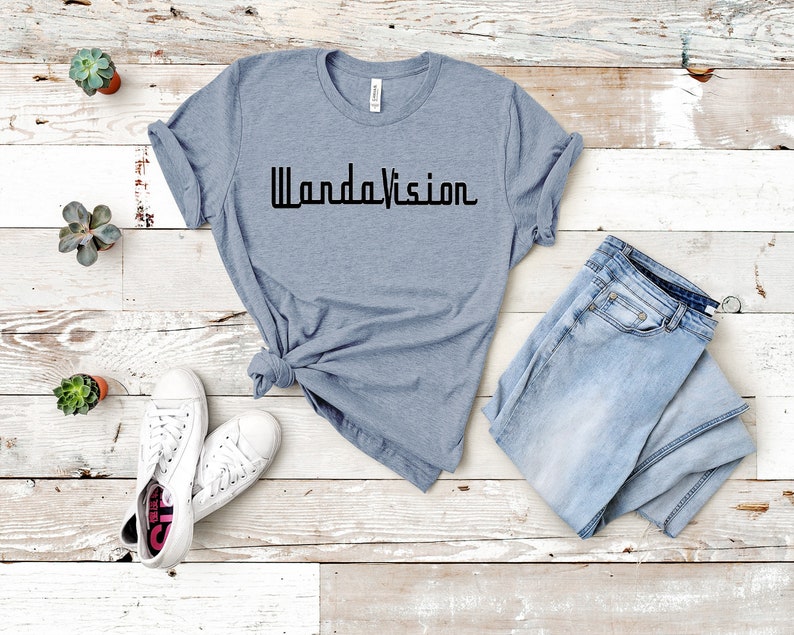 May include: A light blue t-shirt with the word "WandaVision" printed in black. The shirt is paired with light blue jeans and white sneakers. Small potted succulents are arranged around the clothing.