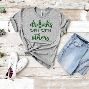 May include: A heather gray t-shirt with the text "drinks well with others" in green. The shirt is paired with light blue jeans and white sneakers. Small potted succulents are arranged around the clothing.