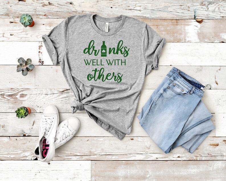 May include: A heather gray t-shirt with the text "drinks well with others" in green. The shirt is paired with light blue jeans and white sneakers. Small potted succulents are arranged around the clothing.
