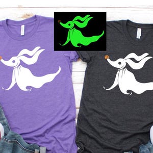 May include: Two t-shirts, one purple and one charcoal gray, each featuring a white graphic of a ghost dog with a pumpkin nose. The shirts are paired with jeans and white sneakers, suggesting casual wear.