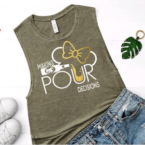 May include: Olive green sleeveless shirt with the text "MAKING POUR DECISIONS" in white. The design includes a wine bottle and a bow. Red sunglasses, white sneakers, and denim shorts are also visible.