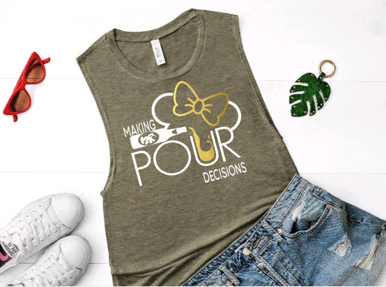 May include: Olive green sleeveless shirt with the text "MAKING POUR DECISIONS" in white. The design includes a wine bottle and a bow. Red sunglasses, white sneakers, and denim shorts are also visible.