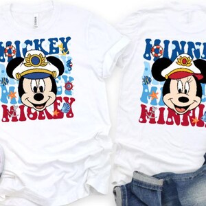 May include: Two white t-shirts featuring Mickey and Minnie Mouse designs. Mickey is wearing a captain's hat, and the text "Mickey" is in red. Minnie is wearing a captain's hat, and the text "Minnie" is in red. The shirts are next to white trainers and blue jeans.