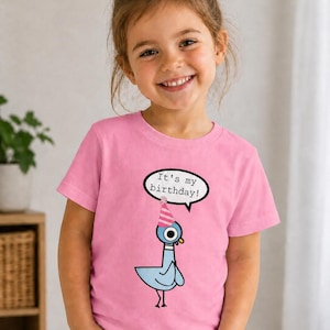 May include: A young girl wearing a pink t-shirt with a cartoon pigeon graphic. The pigeon wears a party hat and a speech bubble says "It's my birthday!". The girl is smiling and looking at the camera.