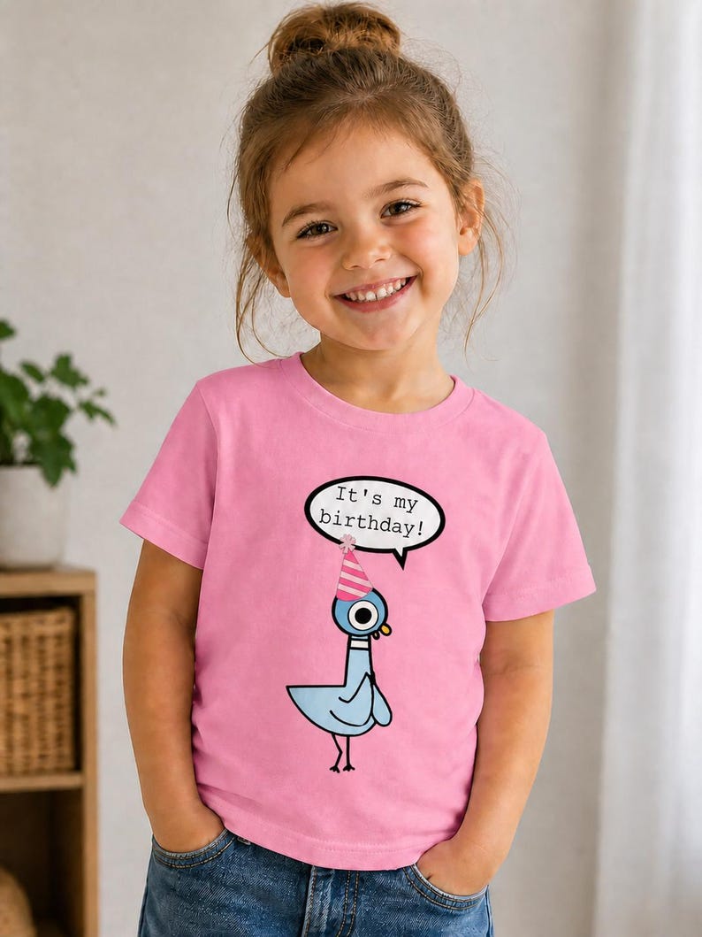 May include: A young girl wearing a pink t-shirt with a cartoon pigeon graphic. The pigeon wears a party hat and a speech bubble says "It's my birthday!". The girl is smiling and looking at the camera.