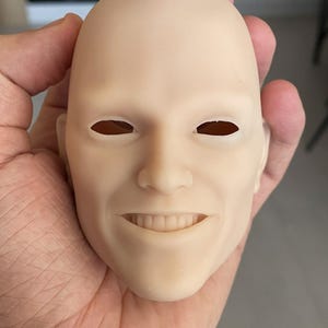 May include: A close-up of a realistic doll head with painted eyes and a smiling mouth. The head is made of a smooth, light-colored material.