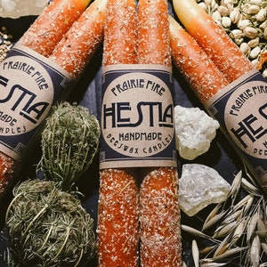 May include: Orange and cream-colored taper candles with a textured, speckled appearance. The candles are bundled together with a label that reads "PRAIRIE FIRE HESTIA HANDMADE BEESWAX CANDLES". The candles are surrounded by dried plants and crystals.