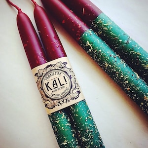 May include: Four hand-dipped beeswax candles. The top portion is a deep burgundy, transitioning to a vibrant green base adorned with dried herbs. The label reads "Prairie Fire Kali! Hand Dipped Beeswax Candles."