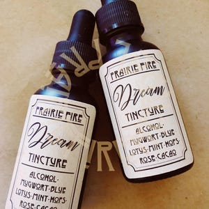 May include: Two dark glass tincture bottles with black droppers. Each bottle has a label that reads "PRAIRIE FIRE Dream TINCTURE" with a list of ingredients: Alcohol, Mugwort-Blue, Lotus-Mint-Hops, and Rose-Cacao.
