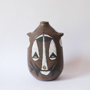Puede incluir: Un jarrón de cerámica marrón con un diseño geométrico blanco y negro. El jarrón tiene un diseño con forma de cara con ojos, nariz y boca.
