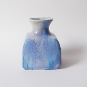 May include: A ceramic vase with a square base and a blue and white glaze. The vase has a narrow neck and a rounded body. The blue glaze fades from a darker tone at the base to a lighter tone at the top. The vase is set against a white backdrop.