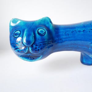 May include: A striking blue ceramic cat ornament with a glossy glaze. The cat features a simplified design with rounded features, including large eyes and a smiling mouth. The ornament is a decorative item.