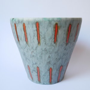 Op de afbeelding: Een keramische vaas met een uitlopende vorm, met een gevlekte blauwgrijze glazuur. Verticale oranje strepen zijn gelijkmatig verdeeld over de buitenkant. De rand heeft een subtiele groene tint. Deze decoratieve vaas is geschikt voor woondecoratie.
