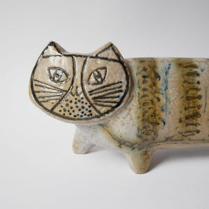 Könnte beinhalten: Keramik-Katzenfigur in Creme- und Beigetönen. Das Gesicht der Katze ist mit schwarzen Details für Augen, Nase und Schnurrhaare versehen. Der Körper hat vertikale Streifen und wird von vier kleinen Beinen getragen. Ein einzigartiges Dekorationsstück.