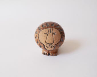 May include: A ceramic lion figurine with a tan face and a brown mane. The lion has a simple, stylized design with a detailed face and a textured mane. The figurine is standing upright on four small legs.