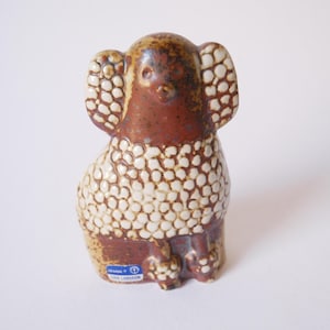 Może przedstawiać: Ceramiczna figurka pudla w kolorze brązowym i białym z fakturowaną powierzchnią. Figurka stoi na białym tle. Figurka jest oznaczona "DESIGN  " i "ULR LARSSON".