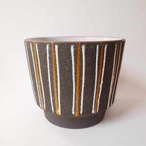 Op de afbeelding: Een donkerbruine keramische plantenpot met verticale witte en oranje strepen. De pot heeft een textuuroppervlak en een licht uitlopende vorm, met een kleine basis. De binnenkant is wit.