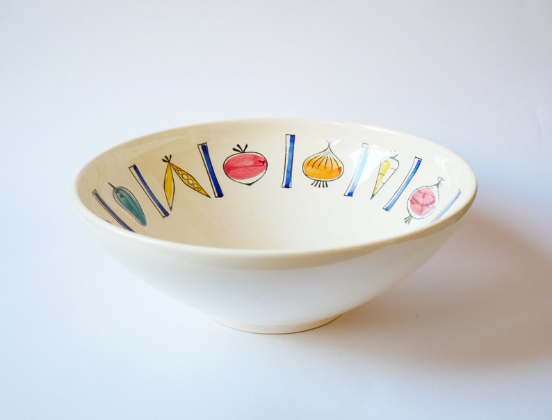 May include: A white ceramic bowl with a blue and yellow vegetable design. The bowl has a wide rim and a rounded bottom.