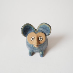 Może przedstawiać: Mała, ręcznie rzeźbiona ceramiczna figurka stworzenia z dużymi, zaokrąglonymi uszami. Korpus pokryty jest niebieskim szkliwem o fakturowanym wykończeniu, natomiast twarz z nieszkliwionej, jasnobrązowej gliny posiada dwoje okrągłych oczu.