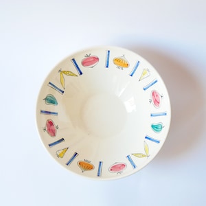 May include: A white ceramic bowl with a blue and yellow rim. The bowl is decorated with a repeating pattern of vegetables, including tomatoes, onions, peppers, and peas.