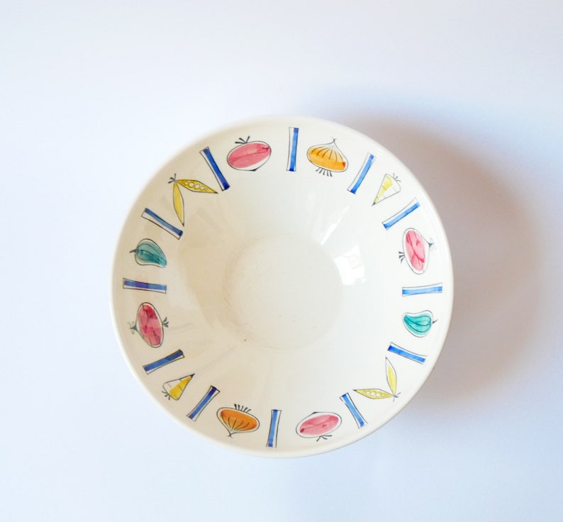 May include: A white ceramic bowl with a blue and yellow rim. The bowl is decorated with a repeating pattern of vegetables, including tomatoes, onions, peppers, and peas.