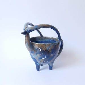 Może przedstawiać: Ręcznie wykonany ceramiczny pojemnik w kształcie ptaka. Niebiesko-brązowa szkliwiona ceramika ma uchwyt i trzy nogi. Białe kropki zdobią korpus. Unikalny przedmiot dekoracyjny. Wymiary w cm.