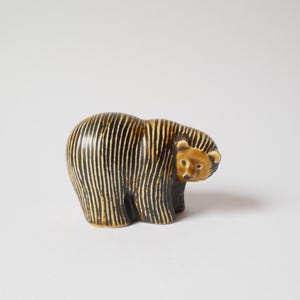 Könnte beinhalten: Eine Keramikbärenfigur mit einem dunkelbraunen Körper und vertikalen cremefarbenen Streifen. Der Bär hat eine glatte, glänzende Oberfläche, eine abgerundete Form und ein einfarbig braunes Gesicht mit kleinen Ohren, dargestellt im Stehen.