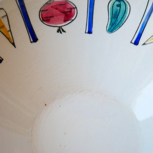 May include: White ceramic bowl with a blue and white rim. The rim is decorated with a repeating pattern of stylized vegetables in red, yellow, green, and blue.