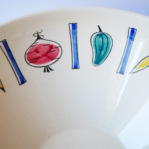 May include: A white ceramic bowl with a blue and yellow vegetable design. The bowl features a carrot, onion, pepper, and pea pods.