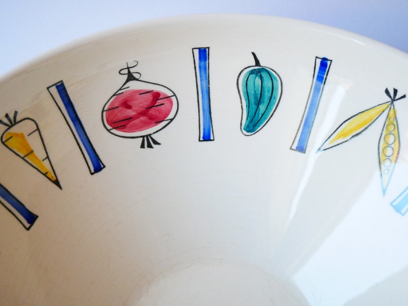 May include: A white ceramic bowl with a blue and yellow vegetable design. The bowl features a carrot, onion, pepper, and pea pods.