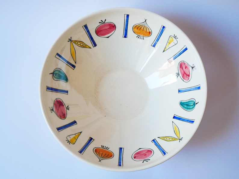 May include: A white ceramic bowl with a blue and yellow rim. The bowl is decorated with a pattern of vegetables, including tomatoes, onions, peppers, and peas.